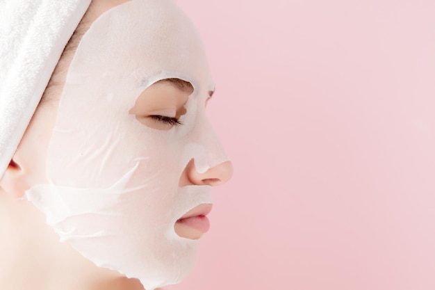 Schöne junge Frau trägt eine kosmetische Gewebemaske auf einem Gesicht auf rosa Hintergrund auf Gesundheits- und Schönheitsbehandlung und Technologiekonzept
