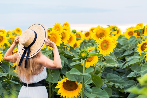 Schöne junge Frau trägt Strohhut und entspannt sich in einem Feld mit Sonnenblumen in einem weißen Kleid