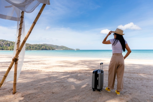 Schöne junge Frau tragen Strohhut mit einem Koffer an einem tropischen Strand