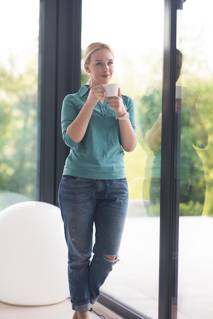 Schöne junge Frau trinkt Morgenkaffee am Fenster in ihrem Haus