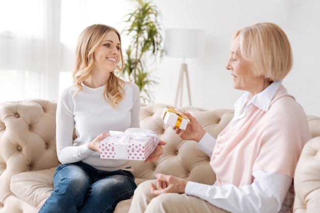 Schöne junge Frau und ihre ältere Mutter sitzen auf dem Sofa und halten Kisten mit Geschenken, während die Tochter ihre Mutter anlächelt