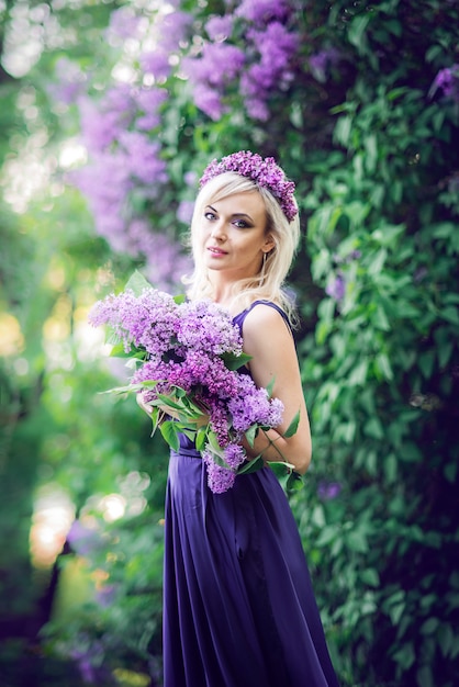 schöne junge Frau von lila Blumen umgeben. Frau sitzt in einem langen Kleid mit einem Schlitz auf dem Hintergrund eines Frühlingsgartens mit Flieder