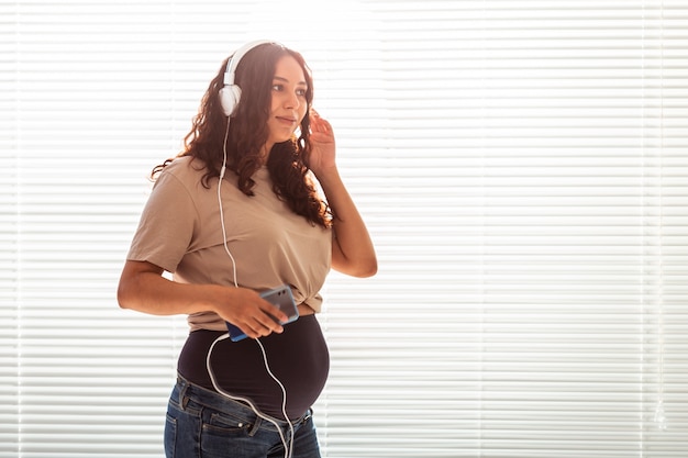 Schöne junge friedliche schwangere Frau hört angenehme klassische Musik mit Smartphone und Kopfhörern. Konzept der positiven Einstellung vor der Geburt