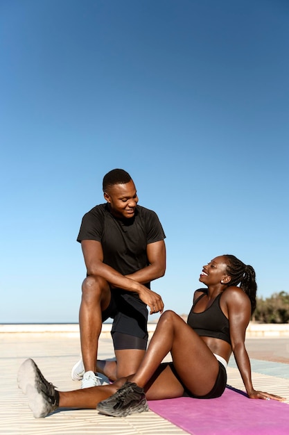 Schöne junge, fröhliche Paare, die sich unterhalten, während sie sich nach dem Training am Strand ausruhen