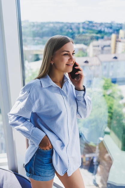 Schöne junge Geschäftsfrau, die auf dem Smartphone spricht und an einem neuen Produktivitätsprojekt für Unternehmen arbeitet, die Freizeit im Coworking Center verbringen und die Fensteransicht genießen