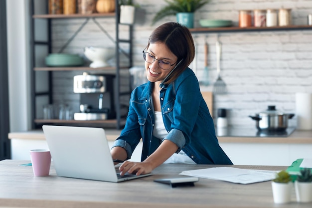 Schöne junge Geschäftsfrau, die mit ihrem Handy spricht, während sie zu Hause mit dem Computer in der Küche arbeitet