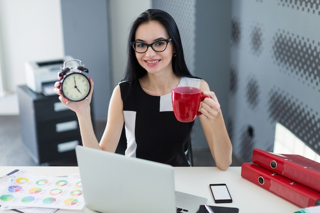 Schöne junge Geschäftsfrau im schwarzen Kleid und in den Gläsern sitzen am Tisch und arbeiten mit Laptop, Griffschale und Wecker