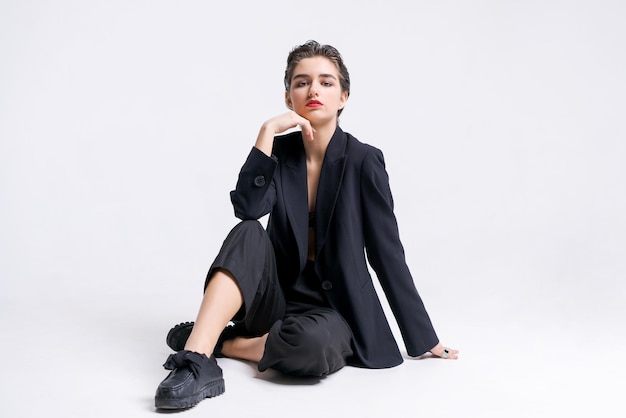 Schöne junge Geschäftsfrau im trendigen schwarzen Anzug posiert auf dem Boden sitzend