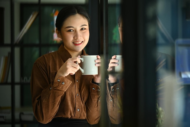 Schöne junge Geschäftsfrau sitzt in der Nähe des Bürofensters und trinkt heißen Kaffee