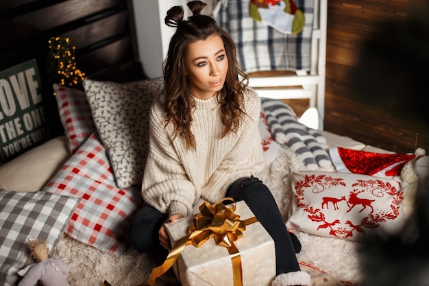 Schöne junge glückliche Frau in einem stilvollen Weinlesepullover mit einem Geschenk auf dem Bett am Heiligabend. Mädchen in einem gestrickten Pullover mit einem Geschenk mit Band auf dem Bett mit Weihnachtsmustern