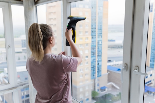 Schöne junge Hausfrau in Arbeitskleidung, die zu Hause Fenster mit professionellen Werkzeugen wäscht.