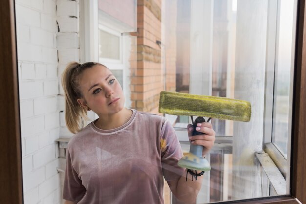 Schöne junge Hausfrau in Arbeitskleidung, die zu Hause Fenster mit professionellen Werkzeugen wäscht.