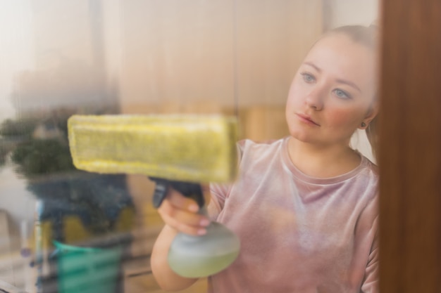 Schöne junge Hausfrau in Arbeitskleidung, die zu Hause Fenster mit professionellen Werkzeugen wäscht.