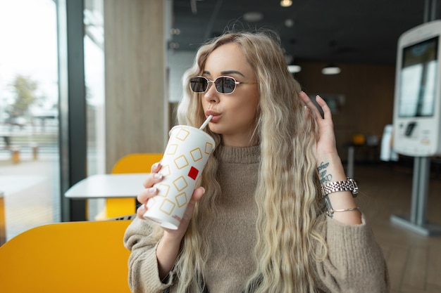 Schöne junge Hipster-Frau mit blonden lockigen Haaren mit Vintage-Sonnenbrille in beigem Pullover hält einen Pappbecher und trinkt Cola in einem Café