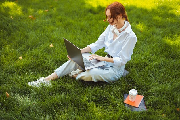 Schöne junge hübsche rothaarige Frau im Park im Freien mit Laptop-Computer für Studium oder Arbeit