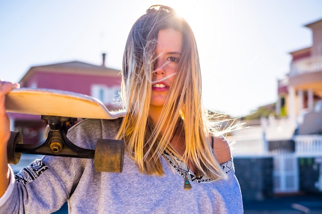 Schöne junge kaukasische Frau, die mit einem Skateboard auf der Schulter, blonde Haare und Sonne in der Hintergrundbeleuchtung, in die Kamera blickt, beherbergt ein defokussiertes alternatives Jugendkonzept in der Stadt