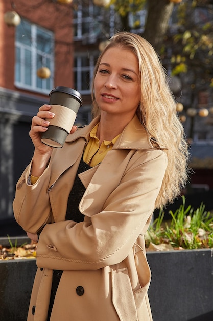 Schöne junge kaukasische lächelnde Frau im Trenchcoat, die draußen steht und eine Tasse Kaffee trinkt