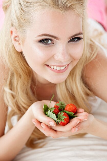 Schöne junge lächelnde Frau, die auf weißem Bett sitzt und einen Haufen wilde Beeren Erdbeeren hält