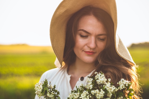 Schöne junge lächelnde Frau im Weinlesekleid und im Strohhut in Feldwildblumen. Das Mädchen hält einen Korb mit Blumen