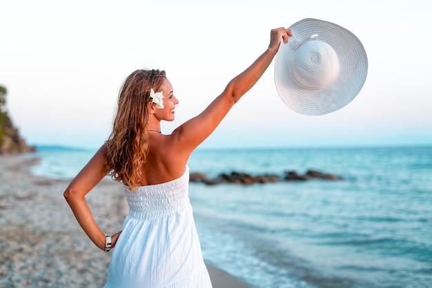 Schöne junge lächelnde Frau im weißen Kleid, die auf den Strand geht und weißen Sommerhut hält.