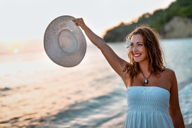 Schöne junge lächelnde Frau im weißen Kleid, die auf den Strand geht und weißen Sommerhut hält.