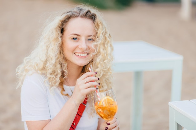 Schöne junge lächelnde frau mit lockigem hellem haar, zahnigem lächeln, erholt sich am strand, trinkt frischen sommercocktail, gekleidet in lässiges weißes t-shirt, hat gute erholung. Menschen, Sommer, Lifestyle-Konzept