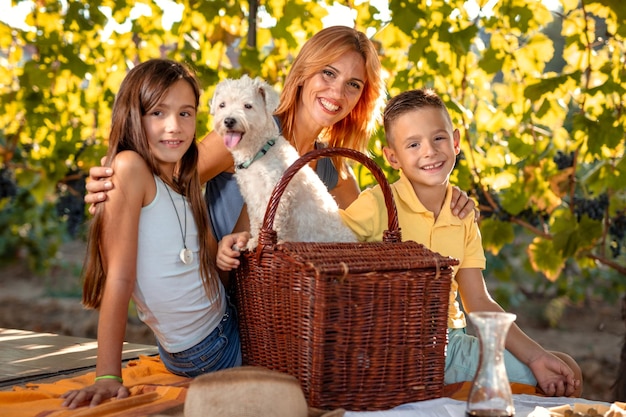 Schöne junge lächelnde Mutter mit ihren Kindern und ihrem Hund beim Picknick auf einem Weinberg.