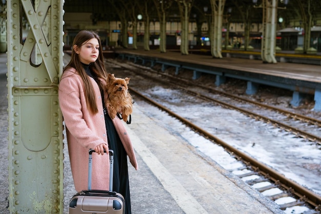 Schöne junge lässige Touristenfrau mit Hund und Koffer, die auf den Zug am Bahnhof wartet, reisen...