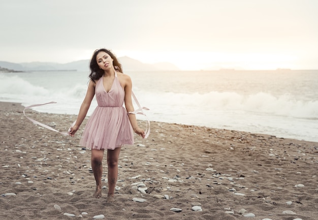 Schöne junge Latina mit langen schwarzen Haaren, die bei Sonnenuntergang am Strand entlang läuft, ein elegantes rosa Kleid trägt, mit Bändern in der Hand spielt und tanzt