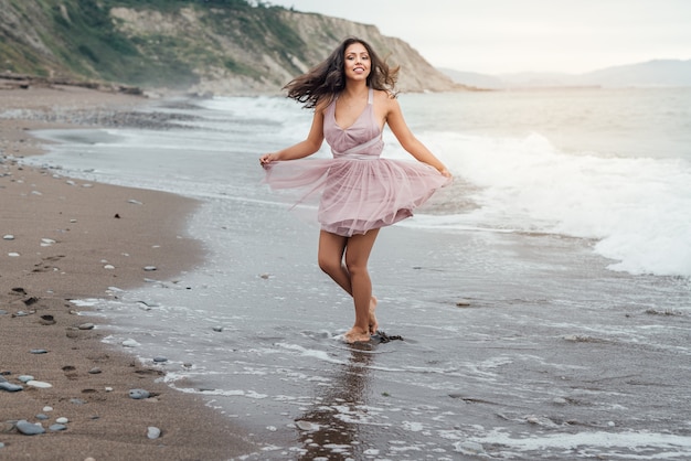 Schöne junge Latina mit langen schwarzen Haaren, die bei Sonnenuntergang am Strand entlang läuft und ein elegantes rosa Kleid trägt