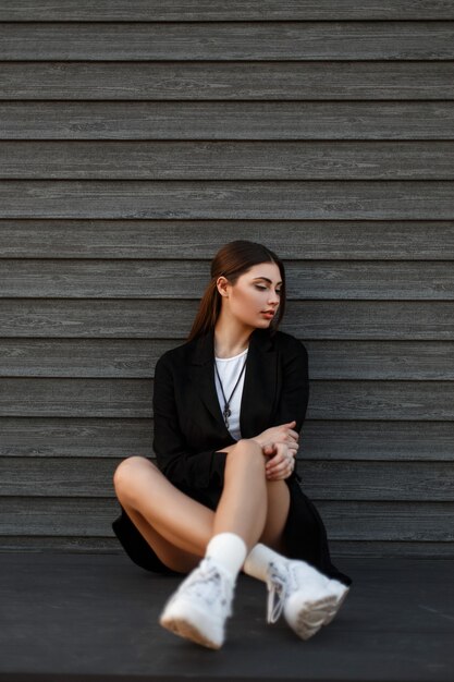 Schöne junge Modellfrau in Mode schwarzen Mantel mit weißen Turnschuhen sitzt in der Nähe einer hölzernen Vintage Wand