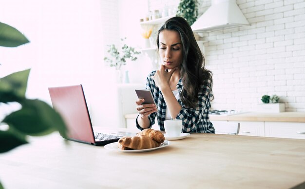 Schöne junge moderne zufällige Frau benutzt Laptop und Telefon in der Küche zu Hause. Online arbeiten. Freiberufler