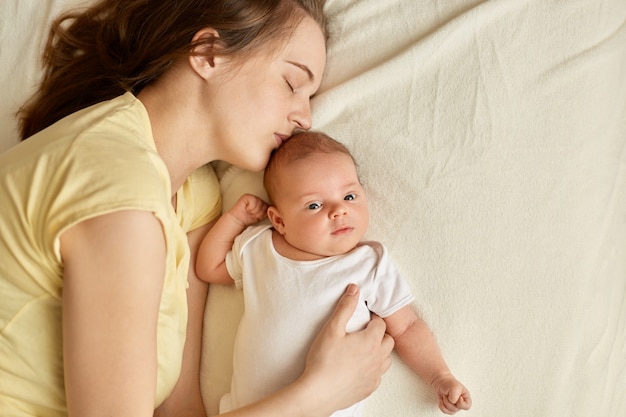 Schöne junge müde schlafende mutter nahe baby, das auf bett auf weißer decke liegt, säugling schaut auf kamera, mama, die ihre tochter oder sohn küsst. mutterschaft.