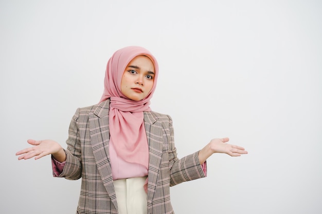 Schöne junge muslimische Geschäftsfrau mit rosa Hijab, die Schultern zuckt, verwirrt aussieht, hat keine Ahnung, steht befragt und unbewusst, ich weiß nicht, warum sie Hände ausbreitet, isoliert auf weißem Hintergrund.