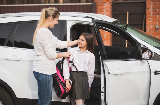 Schöne junge Mutter hilft Tochter, aus dem Auto auszusteigen und die Schultasche anzuziehen
