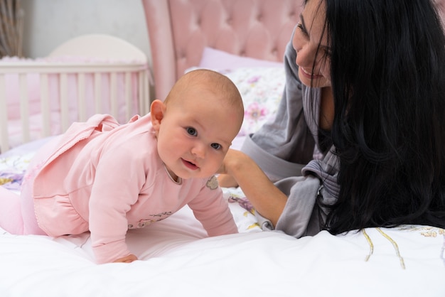 Schöne junge Mutter im Bademantel, die mit ihrem niedlichen glücklichen Baby im rosa Kleid im Bett liegt, das Zeit zusammen genießt und lächelt