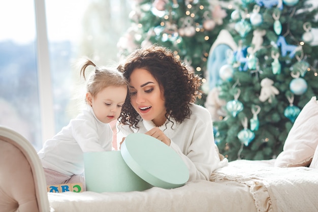 Schöne junge Mutter mit ihrer niedlichen kleinen Tochter, die Weihnachtsferien feiert. Glückliche Familie, die Weihnachtsgeschenke öffnet