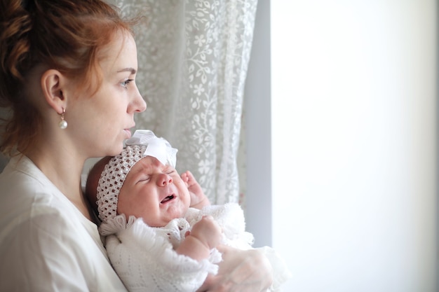 Schöne junge Mutter und neugeborenes Baby in ihren Armen, die am Fenster stehen