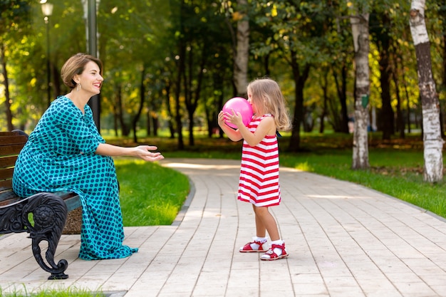 Schöne junge Mutter und Tochter in warmen sonnigen Sommertag. Glückliche Familie Mutter und Kind kleine Tochter spielen mit einem Ballon und spazieren im Park und genießen die schöne Natur.