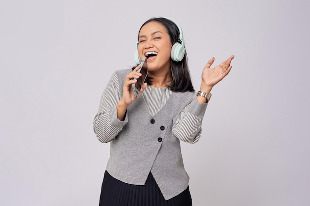 Foto schöne junge sängerin, asiatische frau in den zwanzigern, in einem formellen grauen hemd, die musik in kopfhörern hört und lieder singt, während sie ihre stimme mit einem mobilen telefon aufnimmt, isoliert auf grauem hintergrund