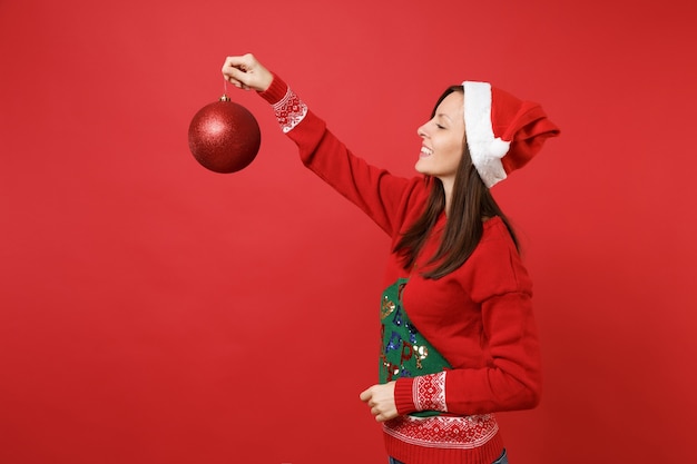 Foto schöne junge santa mädchen in strickpullover weihnachtsmütze auf der suche auf großen glänzenden baum spielzeug in der hand isoliert auf rotem hintergrund. frohes neues jahr 2019 feier urlaub party konzept. kopieren sie platz.