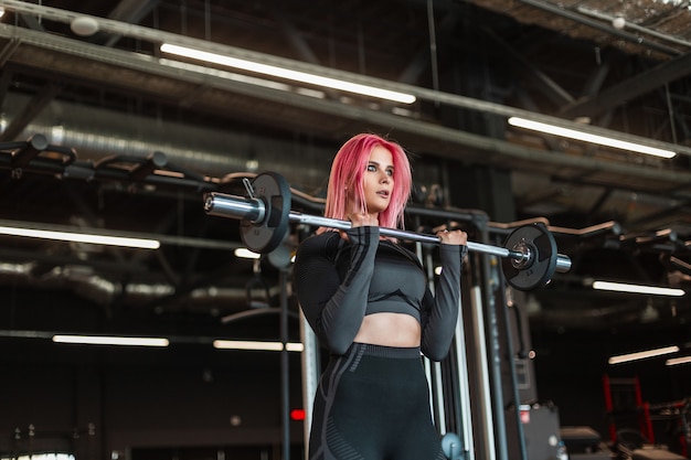 Schöne junge schlanke Frau in schwarzer modischer Sportbekleidung, die mit einer Langhantel und Platten im Fitnessstudio trainiert