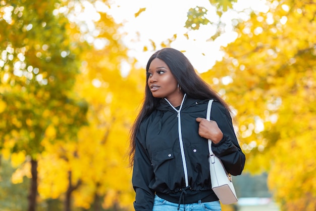 Schöne junge schwarzafrikanische Frau in einer modischen Jacke mit Handtasche geht in einem kanadischen Herbstpark mit leuchtend gelbem Laub