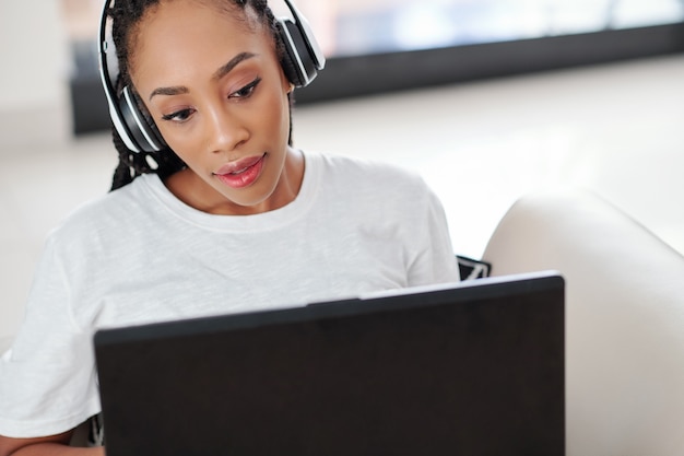 Schöne junge schwarze Frau mit Kopfhörern beim Arbeiten am Laptop zu Hause