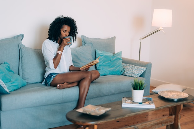 Schöne junge schwarze Frauensitzplätze im Sofa ein Buch lesend und Kaffee trinkend