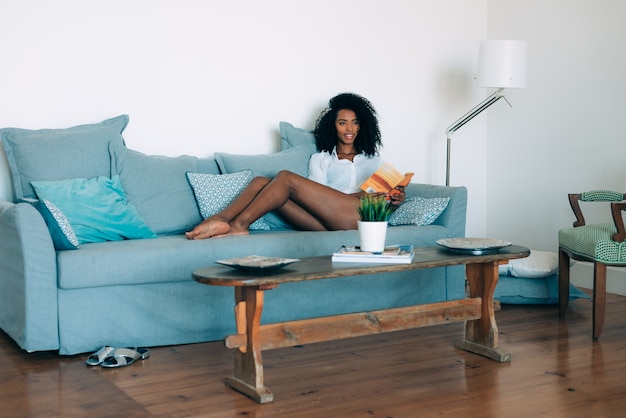 Schöne junge schwarze Frauensitzplätze im Sofa ein Buch lesend