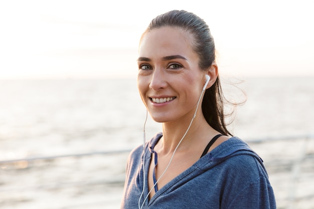 Schöne junge Sportfrau draußen auf der hörenden Musik des Strandes mit Kopfhörern.