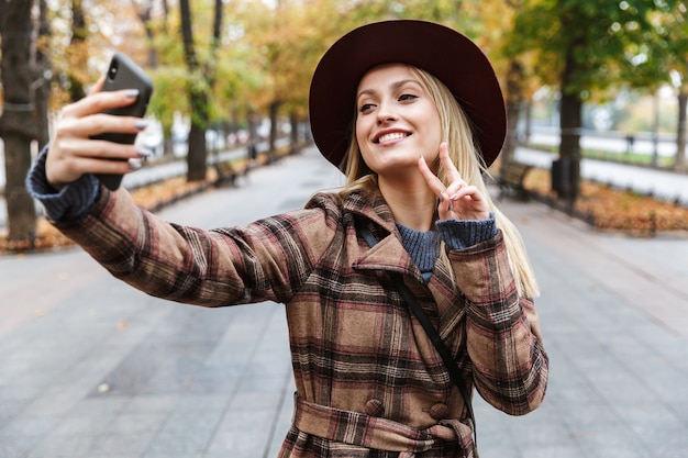 Schöne junge stilvolle blonde Frau, die einen Mantel trägt, der draußen geht und ein Selfie mit Handy nimmt