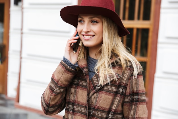 Schöne junge stilvolle blonde Frau, die einen Mantel trägt, der draußen geht, unter Verwendung des Mobiltelefons