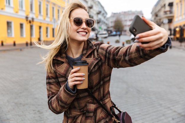 Schöne junge stilvolle blonde Frau, die einen Mantel trägt, der draußen geht